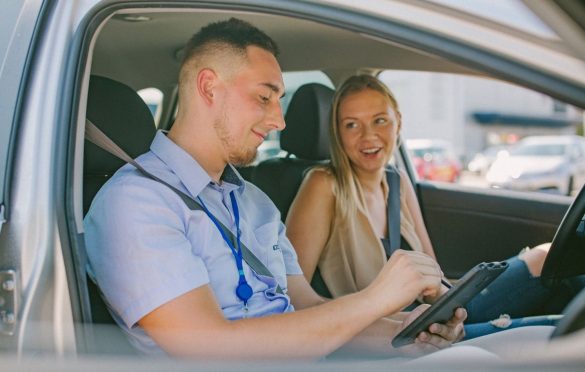 Egzaminuotojas isėdi automobilyje ir žymisi kažką į planšetę, o vairuotojas šypsosi. Abu su diržais.