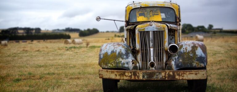 : Senas, surūdijęs sunkvežimis stovi lauke, apsuptas šieno ritinių ir kaimiško peizažo. Matomas nusilupęs dažas, pažeistas priekis ir debesuotas dangus fone.